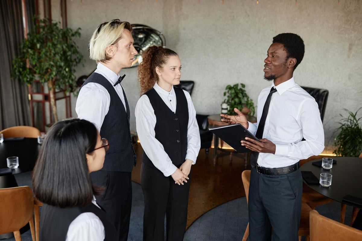 Side view portrait of young Black man as restaurant manager talking to servers wearing classic uniforms during morning instructions