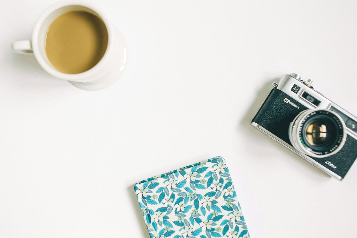black and grey SLR camera, floral notebook, and white ceramic mug