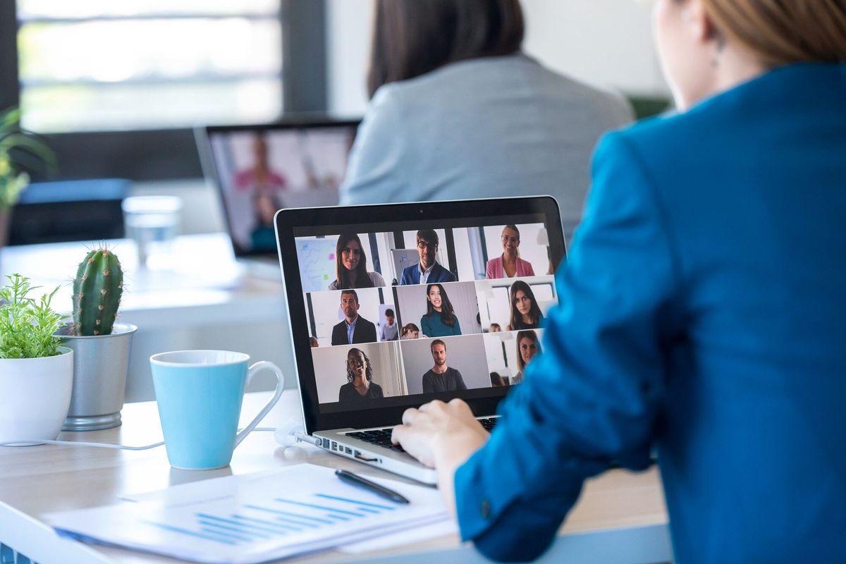Back view of two business women speaking on video call with diverse colleagues on online briefing with laptop in the office.