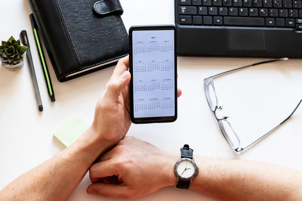 Mobile phone with open calendar for 2022 year in male hand on working desk, laptop, pen, clock, keys