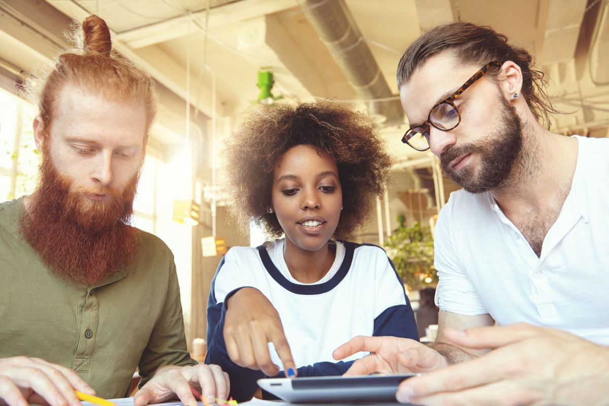 Three creative people having discussion at cafe: African woman explaining her vision, pointing at screen of touch pad, bearded man in glasses listening attentively and redhead partner making notes