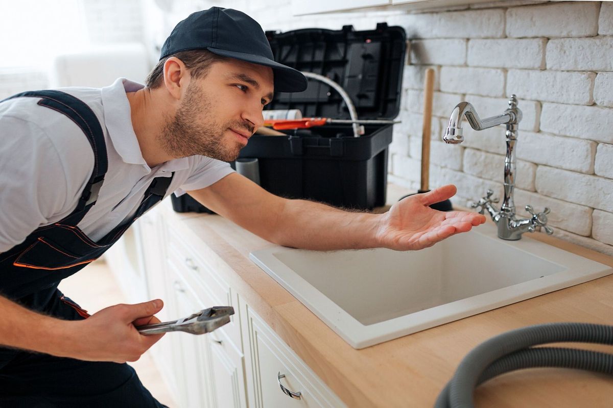 Young handyman check tap for leaking. He hold hand under it. Man stand in kitchen at sink. Toolbox and hose on desk.
